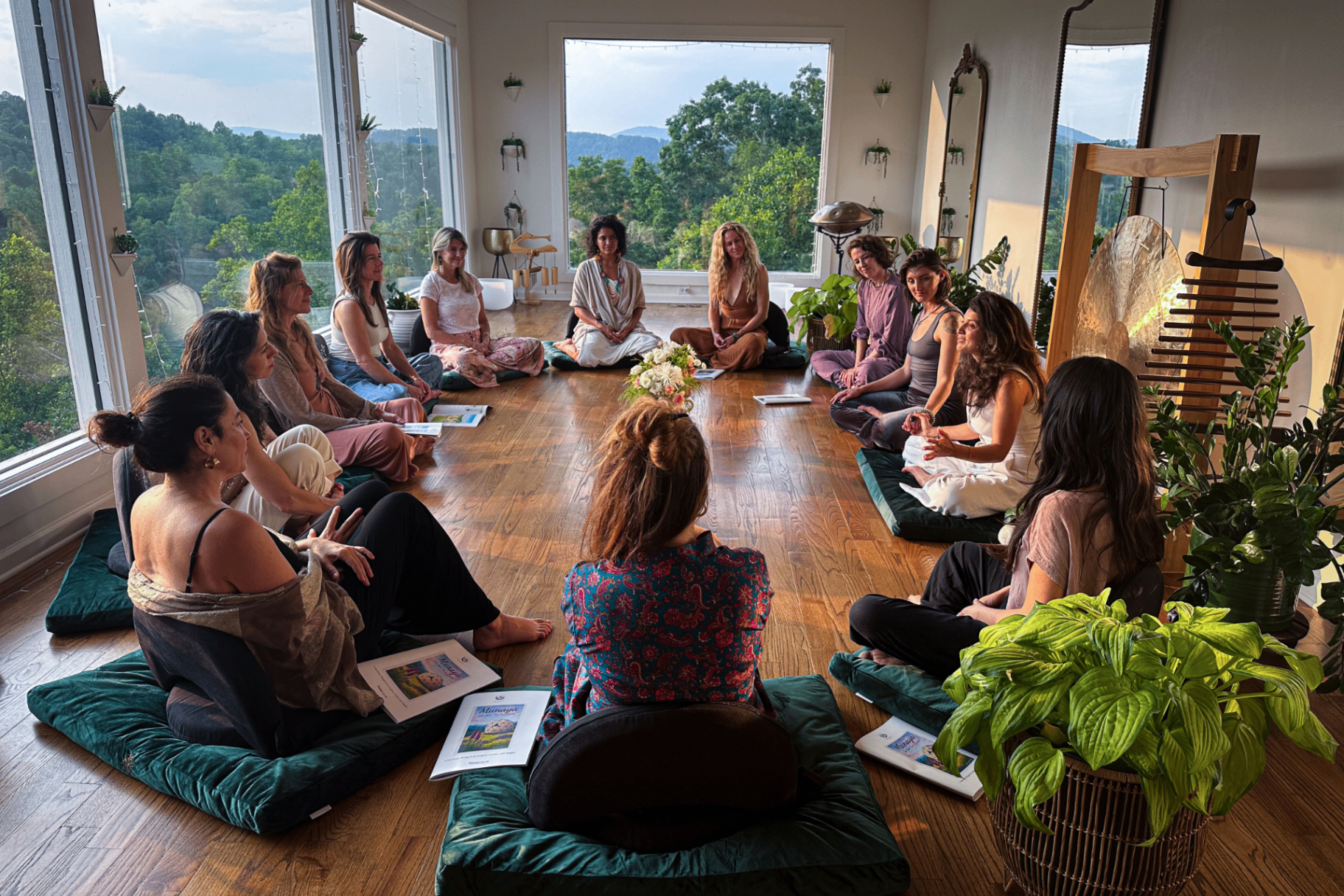 A circle of women seated on green cushions in the Glass Studio, mountains through the windows